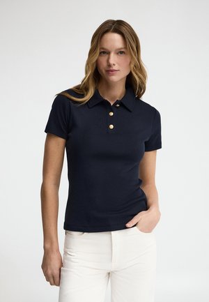 Frau mit welligem hellbraunem Haar trägt ein marineblaues Kurzarm-Poloshirt mit drei goldenen Knöpfen und weiße Hose, steht vor einem schlichten Hintergrund.