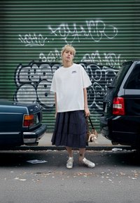 White cotton t-shirt with logo, navy plaid tiered skirt, gray shoes, holding a small bag. Urban setting with graffiti, cars, and sidewalk debris.