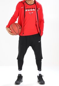 Sudadera roja con cremallera y detalles en negro, camiseta sin mangas roja con el texto y el logotipo de "BULLS", pantalones cortos negros, leggings negros y zapatillas de deporte negras con detalles blancos.