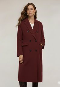 Cappotto bordeaux doppiopetto con colletto a revers, tasche laterali e bottoni neri. Tessuto morbido, design di lunghezza media.