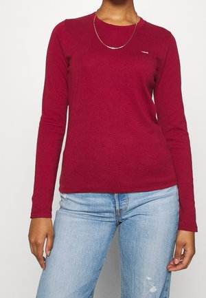 Person wearing a plain maroon long-sleeve shirt and light blue jeans with a thin gold necklace against a plain background.