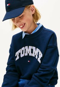 Sudadera azul marino con "TOMMY" bordado en blanco, que presenta un pequeño logotipo de bandera. Debajo, se puede ver una camisa de cuello de rayas azul claro.