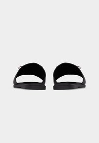 Paire de sandales plates noires à bout ouvert avec une large bride unique sur le pied, vues de face sur un fond clair uni.