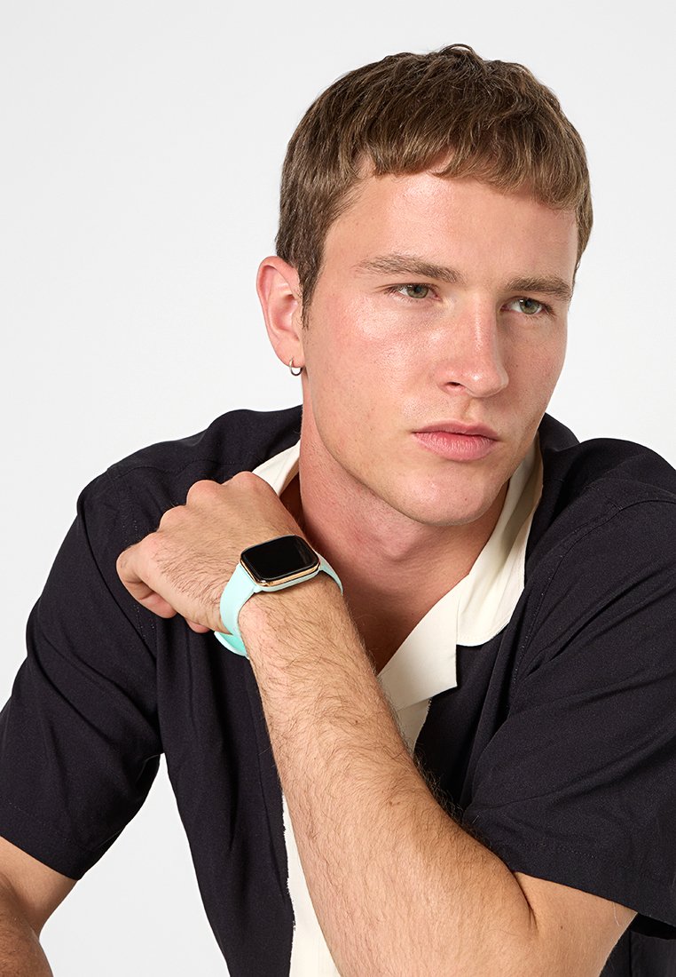 Reloj inteligente con carátula cuadrada, hardware dorado y una correa de silicona azul clara. El sujeto lleva puesta una camisa negra con cuello blanco.