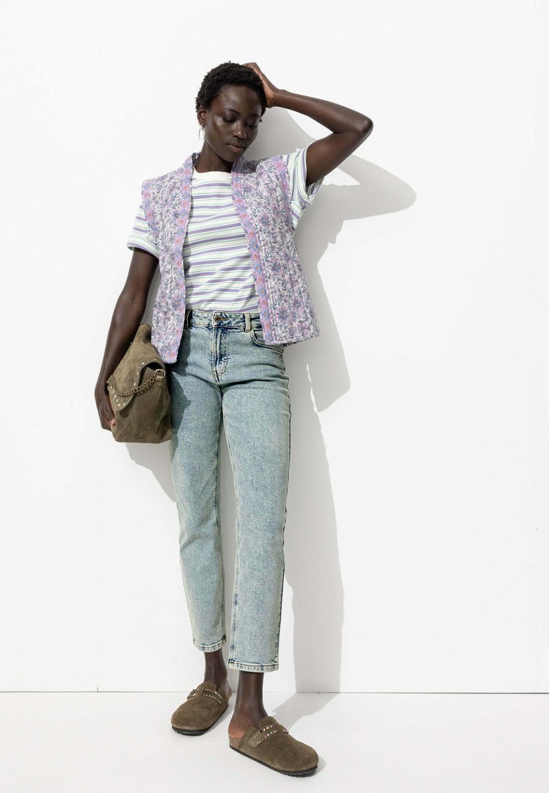 Gilet sans manches à motif floral lavande sur un t-shirt rayé. Jean taille haute bleu clair et sabots marron avec des clous. Sac olive.