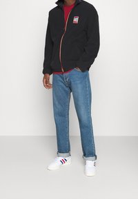 Černá fleecová bunda na zip s vysokým límcem, s barevným logem, v kombinaci s modrými džíny a bílými teniskami s červenými a modrými pruhy.