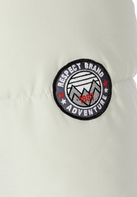 Kreminės spalvos audinys su siuvinėta logotipo etikete, kurioje užrašyta "Gerbk Prekės Nuotykius" ir pavaizduota kalnų tema, akcentuota raudonais žvaigždutėmis.