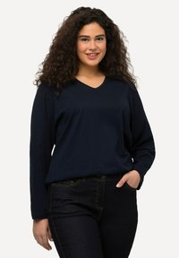 Marineblaues Langarm-V-Ausschnitt-Shirt aus glattem Stoff, kombiniert mit dunklen Jeans. Einfaches Design mit legerer Passform und minimaler Detailgestaltung.