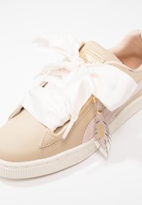 Baskets en cuir beige avec un accent rose clair, lacets satin blancs surdimensionnés, semelle texturée et une étiquette en cuir décorative avec des détails dorés.