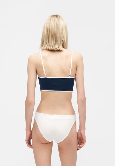Femme aux cheveux blonds courts portant un crop top bleu marine avec des bordures blanches et un sous-vêtement blanc, debout de dos face au spectateur.