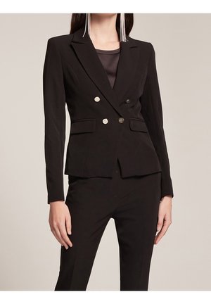Woman wearing tailored black double-breasted blazer with silver buttons, paired with matching fitted trousers and long white tassel earrings.