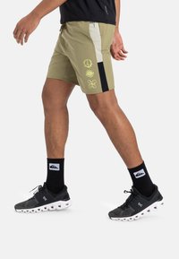 Bermudas verde oliva con paneles negros y beige, que presentan diseños gráficos amarillos estilizados en la parte frontal. Tejido suave, de longitud por encima de la rodilla.