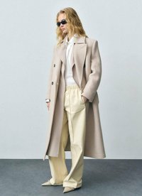 Lichtbeige wollen jas met een notch-kraag, gecombineerd met een wit shirt en crèmekleurige tapered broek. Het model draagt zwarte zonnebril en puntige schoenen.