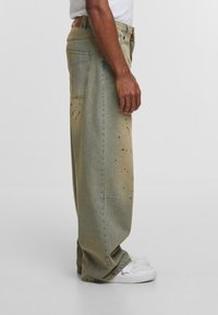 Bokova perspektiva osobe koja nosi široke izblijele traperice s mrljama od boje i bijele tenisice na jednobojnoj pozadini.