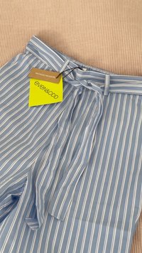 Pantalons rayés bleu clair en polyester recyclé. Présentent une taille à nouer et une étiquette beige avec le nom de la marque et des informations sur le matériau.