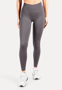 Leggings taille haute, ajustés, en tissu gris foncé, avec une texture lisse sans motifs. Associés à des baskets de sport blanches.
