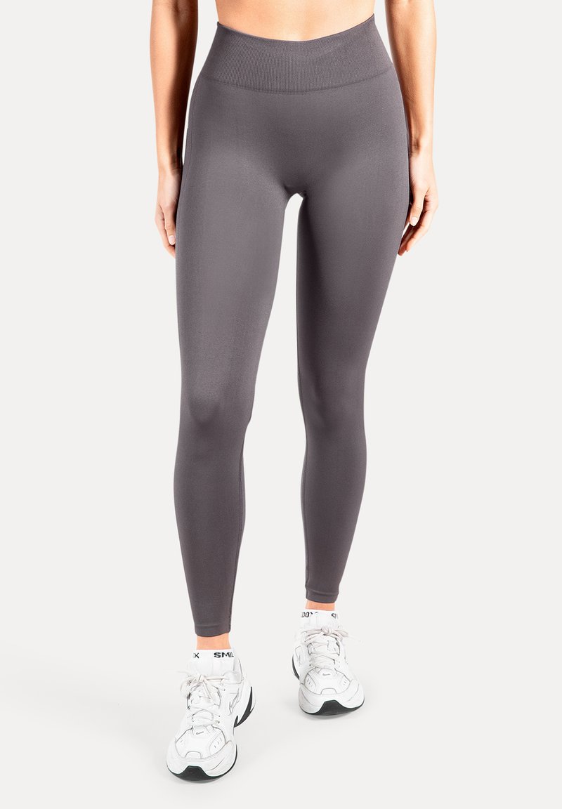 Leggings taille haute, ajustés, en tissu gris foncé, avec une texture lisse sans motifs. Associés à des baskets de sport blanches.