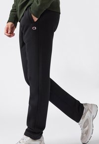 Man in zwarte Champion joggingbroek, groene top en witte sneakers, die met één hand in zijn zak tegen een effen achtergrond loopt.