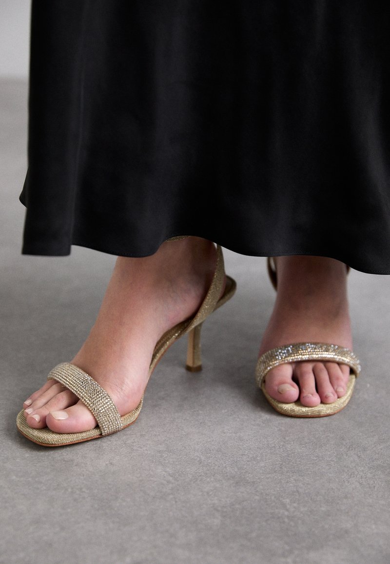 Pieds portant des sandales à talons hauts dorées avec des brides ornées de strass, debout sur un sol gris sous une longue jupe noire.