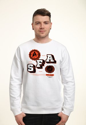 Man die een wit sweatshirt draagt met zwarte en oranje Starfleet Academy-logo's en tekst, staande tegen een effen lichte achtergrond.