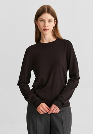 Femme aux cheveux bruns et raides portant un pull noir à manches longues et un pantalon gris foncé, debout les mains jointes devant elle.