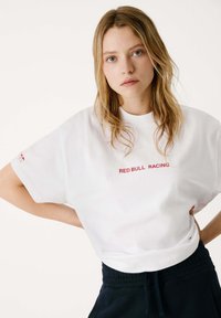 Camiseta de algodón blanca con un corte relajado, que presenta el texto rojo "RED BULL RACING" en la parte frontal y un sutil logotipo en la manga izquierda.