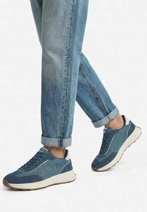 Osoba nosząca podwinięte niebieskie dżinsy i niebieskie trampki marki G-Star z białymi i gumowymi podeszwami, stojąca z jedną lekko uniesioną stopą.