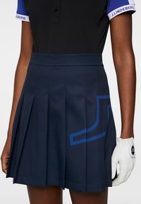 Mörkblå plisserad kjol med en blå geometrisk design, tillverkad av tyg. Åtföljd av en vit golfhandske med en logotyp.