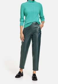 Žena v tyrkysovém žebrovaném svetru, tmavozelených kožených kalhotách a černých mokasínech, stojí s jednou rukou v kapse na bílém pozadí.