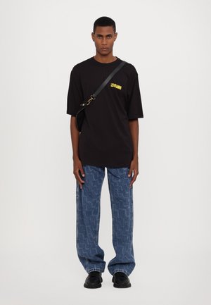 Black oversized t-shirt with yellow logo, paired with blue patterned baggy jeans and black shoes. Simple design, casual fit.