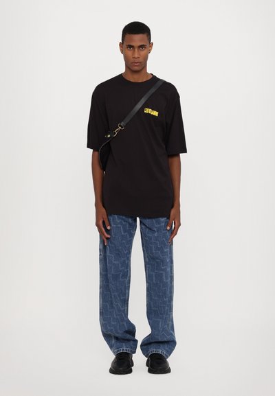 Black oversized t-shirt with yellow logo, paired with blue patterned baggy jeans and black shoes. Simple design, casual fit.