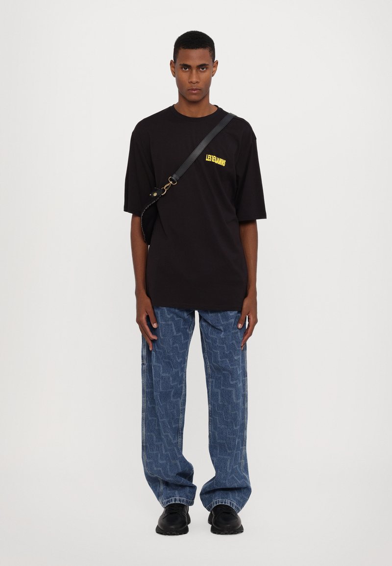 Juodas per didelis t-shirt su geltonu logotipu, derinamas su mėlynomis raštuotomis laisvomis džinsais ir juodais batais. Paprastas dizainas, kasdienis siluetas.