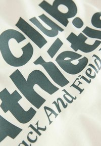 Camiseta de tela cremosa con gran texto en verde oscuro que dice "Club Athletics Track And Field". Textura suave, diseño casual.