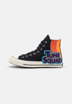 Sort højtop-lærredssneaker med flerfarvede orange og gule accenter og en fremtrædende blå "TUNE SQUAD"-logo. Gummisnude og sål.