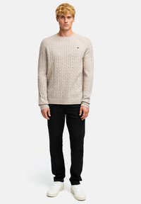 Beżowy sweter z splotem w warkocz, z okrągłym dekoltem, ściągaczami przy mankietach i u dołu. W połączeniu z czarnymi spodniami i białymi trampkami, stojący na jednolitym tle.