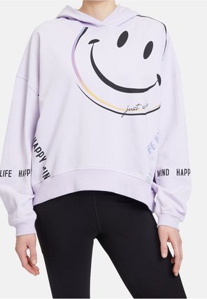 Jasnofioletowa bluza z kapturem z dużą czarną uśmiechniętą buźką, napisem „just smile” i „happy mind happy life” na rękawach, noszona z czarnymi spodniami.