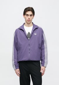 Lilla lukuga jope, mille varrukatel on valge kolmetriibuline disain ja rinnal väike logo. Kerge, sile tekstuur, vabaaja sobivus.