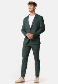 Zielony garnitur o teksturowanej powierzchni z dopasowanym krojem, jednorzędową marynarką zapinaną na jeden guzik oraz wąskimi spodniami, zestawiony z kremowymi butami sportowymi i białym t-shirtem.