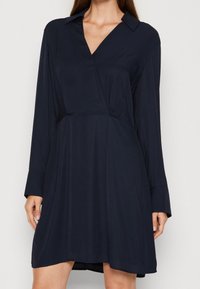 Navyblaues Kleid aus glattem Stoff, mit klassischem Kragen, langen Ärmeln und einer A-Linien-Form mit einem drapierten Vorderteil.