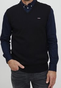 Mann trägt einen schwarzen ärmellosen V-Ausschnitt-Pullover über einem marineblauen Hemd mit Knopfleiste und dunkelgrauen Jeans, Hand in der Tasche.