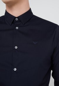 Mörkblå skjorta med knappar gjord av slät tyg, med en klassisk krage, silverfärgade knappar och en diskret broderad logotyp på bröstet.