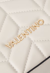 Kvalitatīva krēmkrāsas ādas soma ar izteiktu zelta "VALENTINO" logotipu. Pamanāmas šuves un teksturēta virsma uzlabo dizainu.