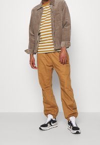 Tan corduroy jakke, stribet gul og hvid bomulds t-shirt, løse tan bukser og sorte Nike sneakers med hvide detaljer.