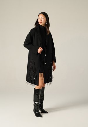 Cappotto in lana nera con vestibilità rilassata, bottoni bianchi e dettagli in frange con perline lungo l'orlo. Abbinato a stivali alti neri lucidi fino al ginocchio.