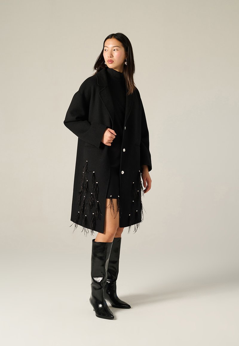 Cappotto in lana nera con vestibilità rilassata, bottoni bianchi e dettagli in frange con perline lungo l'orlo. Abbinato a stivali alti neri lucidi fino al ginocchio.