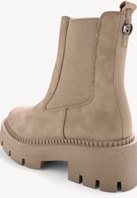 Beige ankle boot with a textured fabric panel, elastic sides, chunky rubber sole, and a circular metal accent near the heel.