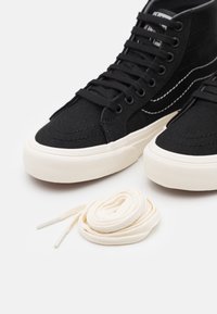 Černé plátěné sneakers s vysokým vrškem mají bílé švy, krémové gumové podrážky a černé tkaničky. Obsahuje také další pár krémových tkaniček.