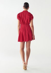 Rotes kurzes Kleid mit Puffärmeln, elastischem Bund, schwingendem Rock und Ausschnittrücken. Das Model trägt hautfarbene High-Heels.