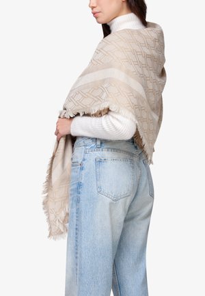Sciarpa beige con motivo a frange, indossata sopra un maglione bianco, che si drappeggia elegantemente intorno alle spalle, abbinata a jeans azzurri chiari.