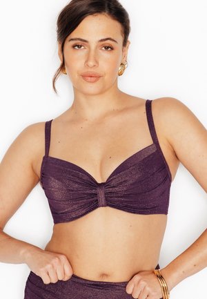 Donna con capelli scuri, indossa un reggiseno viola scintillante e slip abbinati, con orecchini a cerchio dorati e braccialetti d'oro, posa su sfondo bianco.
